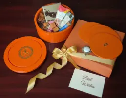 Luxurious orange Chocolics gift hamper featuring a reusable wooden box filled with assorted chocolates, chocolate-coated nuts, cookies, flavoured tea, almonds, nut bites, satin ribbon packaging, 5 placemats, and a personalised note card displayed on a wooden surface.
