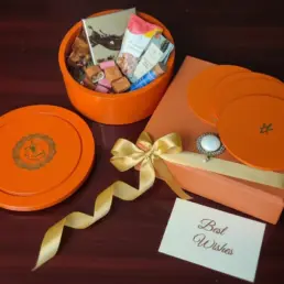 Chocolics Royal Signature Luxury Hamper 10 Luxurious orange Chocolics gift hamper featuring a reusable wooden box filled with assorted chocolates, chocolate-coated nuts, cookies, flavoured tea, almonds, nut bites, satin ribbon packaging, 5 placemats, and a personalised note card displayed on a wooden surface.